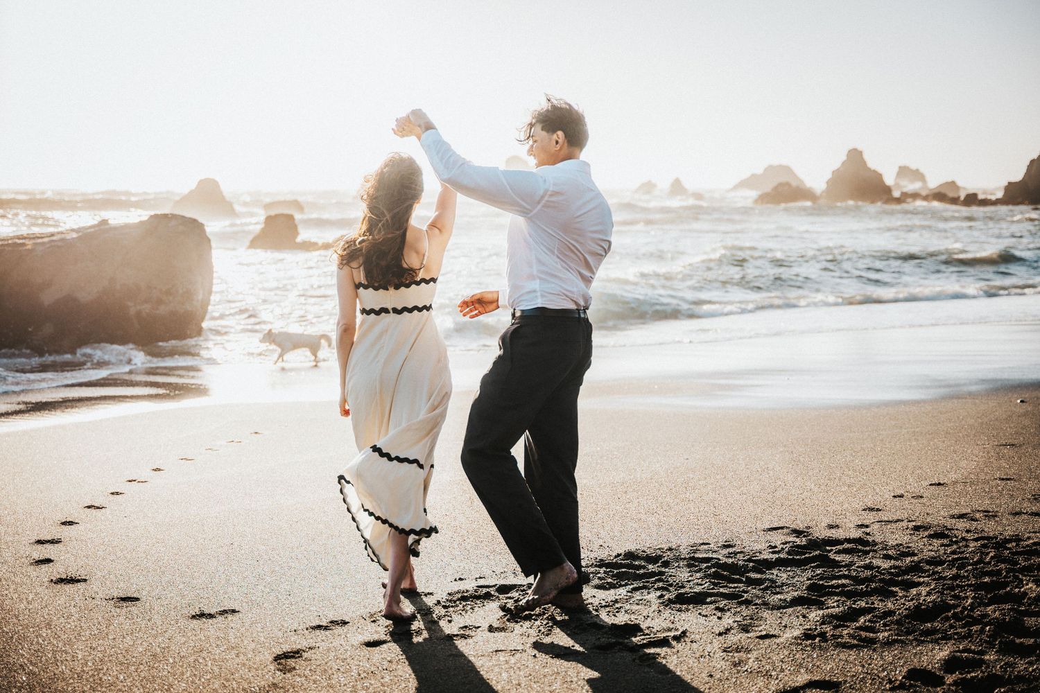 Couple dancing on a sandy beach at sunset, woman in white dress being twirled by man in white shirt.