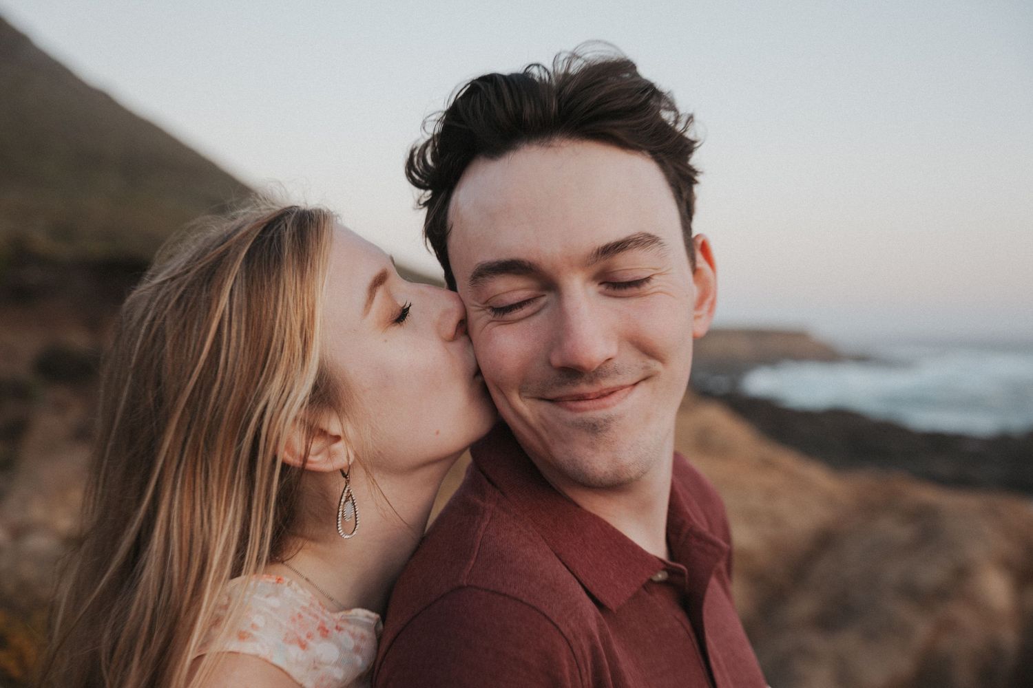 A woman kisses a smiling man on the cheek outdoors with a scenic coastal hillside background.