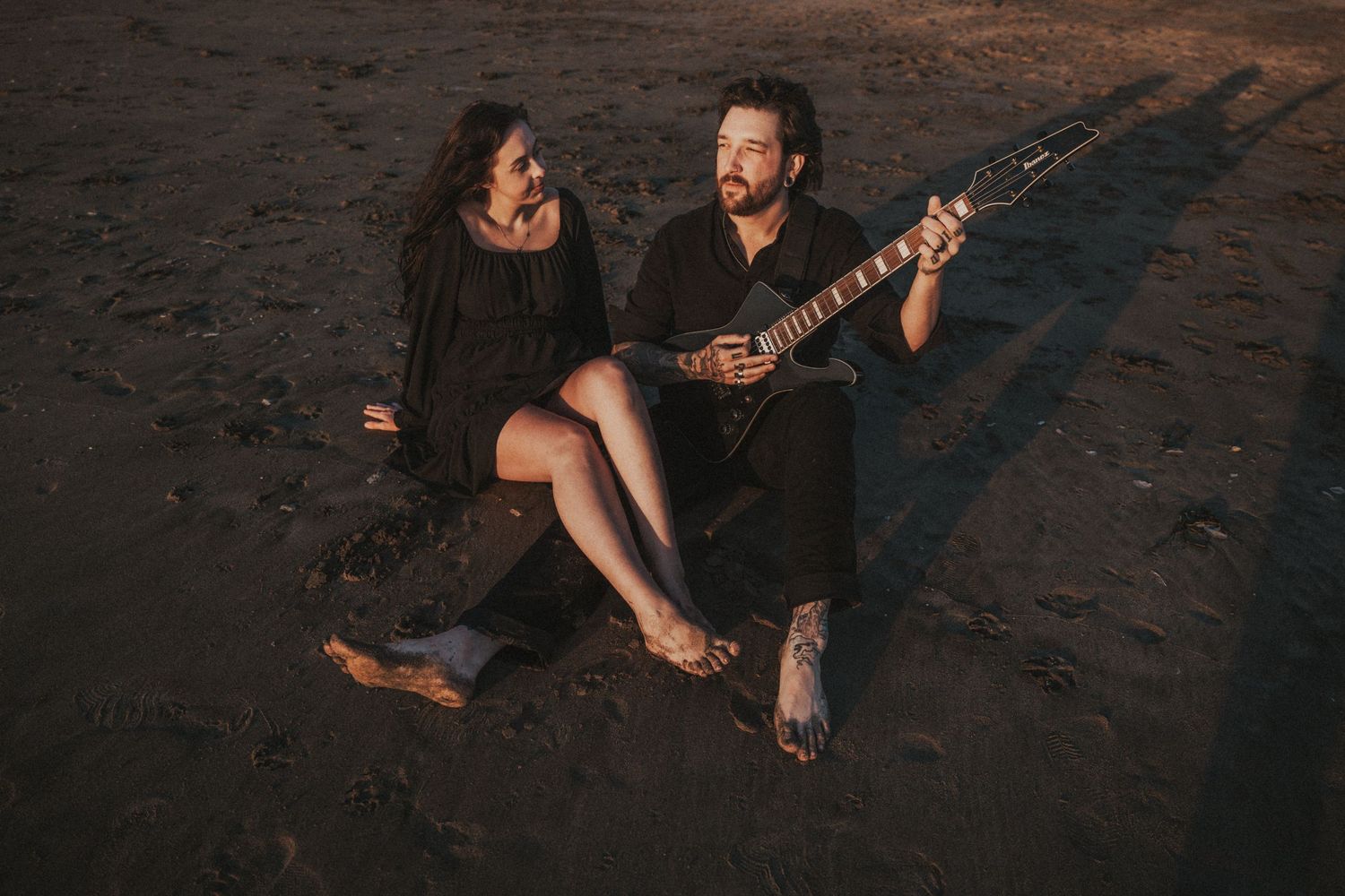 Couple sitting on beach at sunset, man playing electric guitar, woman in black dress smiling at him.