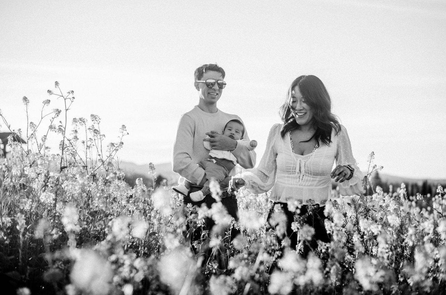 Black and white photo of couple walking through wildflower field while carrying their baby.