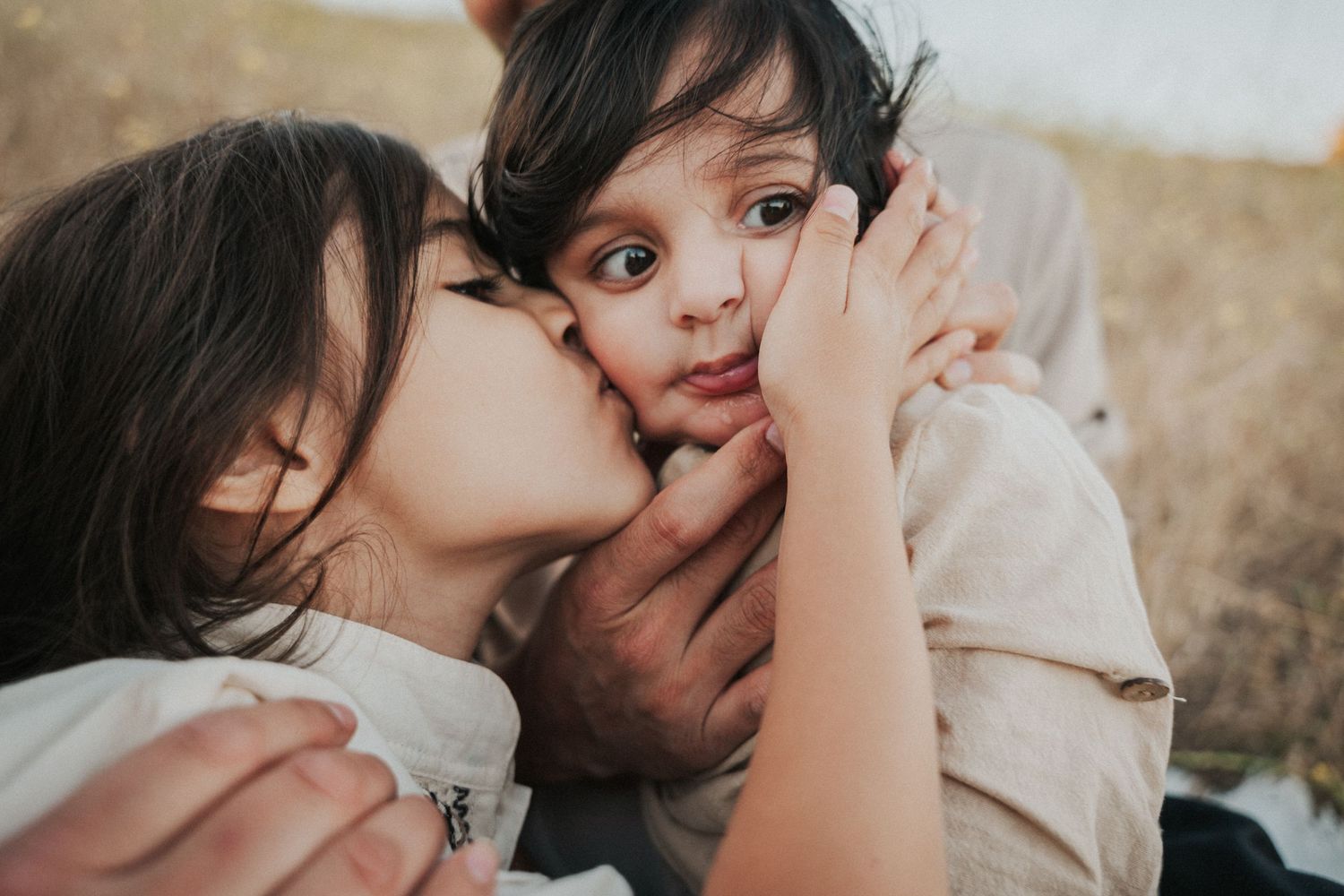 A young girl tenderly kisses a toddler boy on the cheek while an adult holds him in a warm family moment.