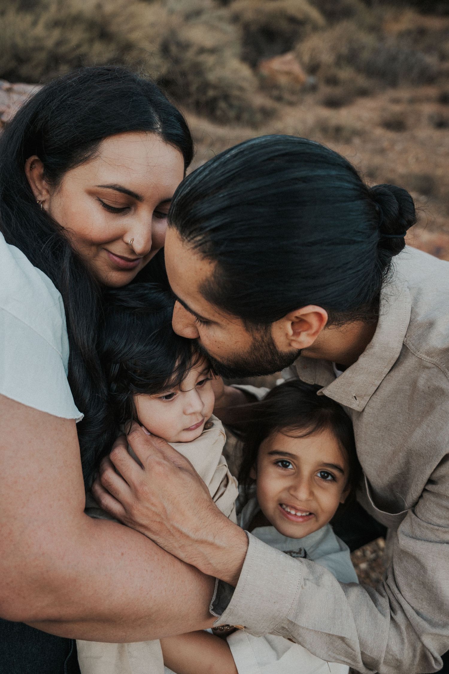 A loving family of four embraces outdoors, with parents hugging two young children in a warm, candid moment.