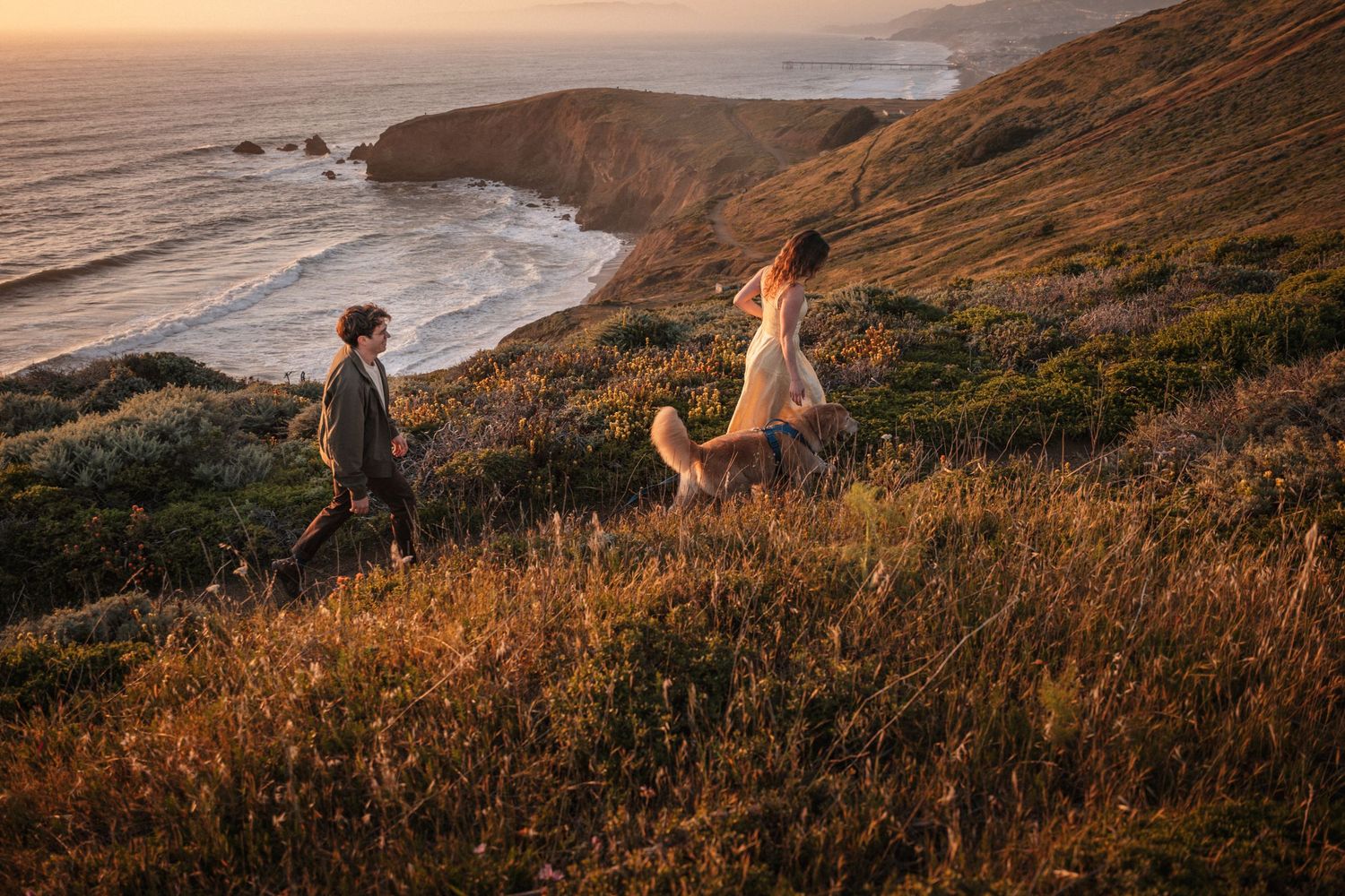 Couple running through golden coastal cliffs at sunset, with ocean waves and rocky shoreline in the background.