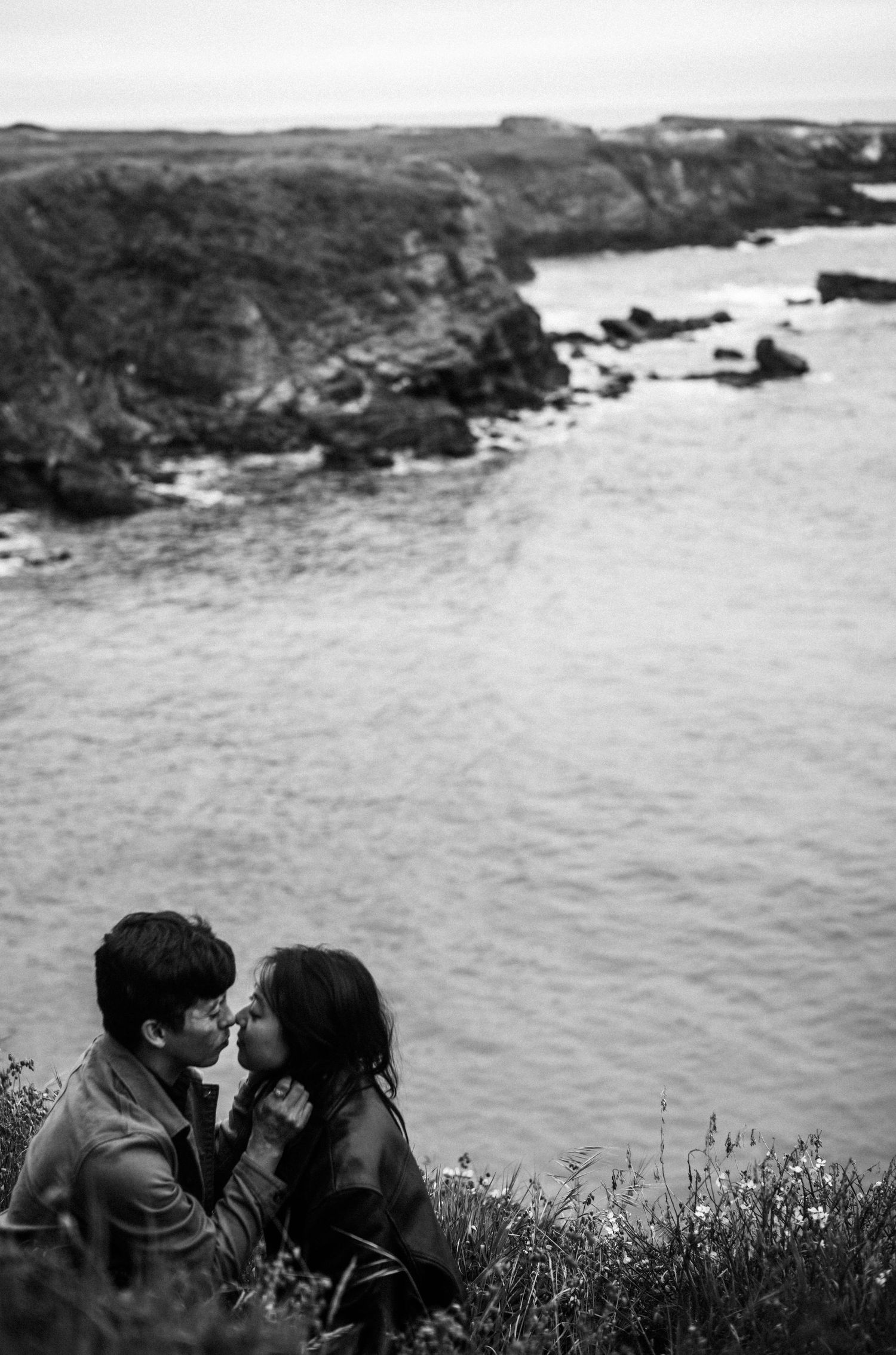 Couple sharing a romantic kiss on a coastal cliff overlooking dramatic rocky shoreline in black and white photo.