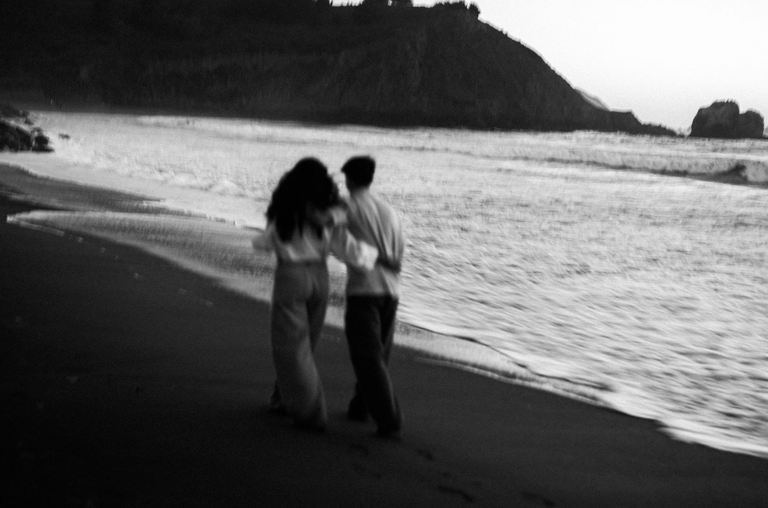 Couple walking hand in hand along a black sand beach at dusk in black and white photography.