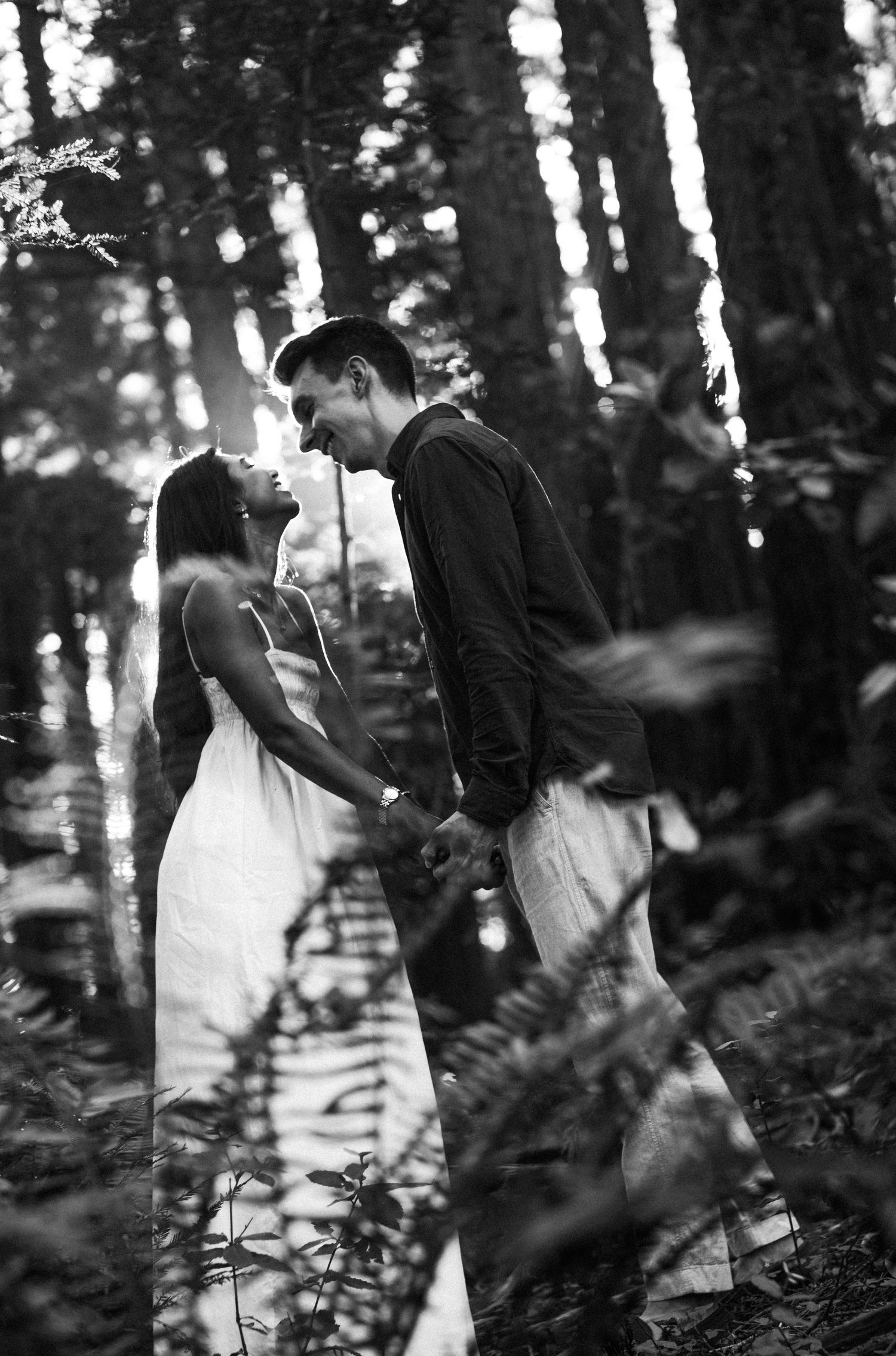 Black and white photo of couple holding hands and gazing at each other among tall trees in a forest.