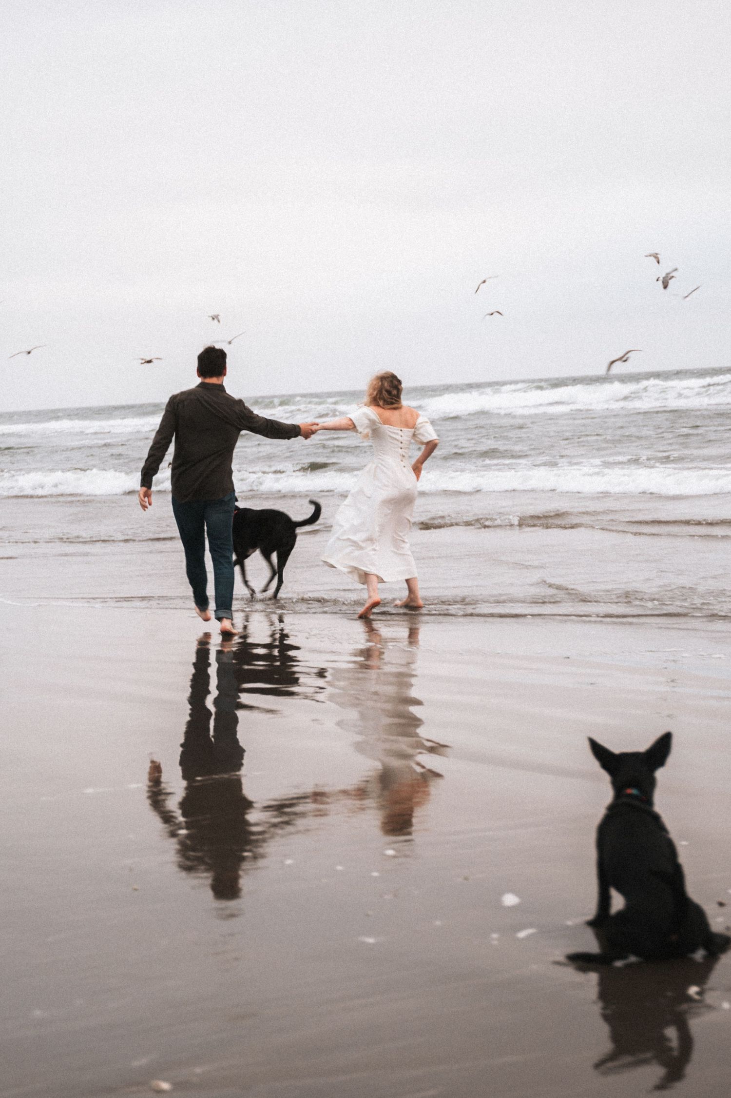 Couple holding hands running into ocean waves with two black dogs on cloudy beach.