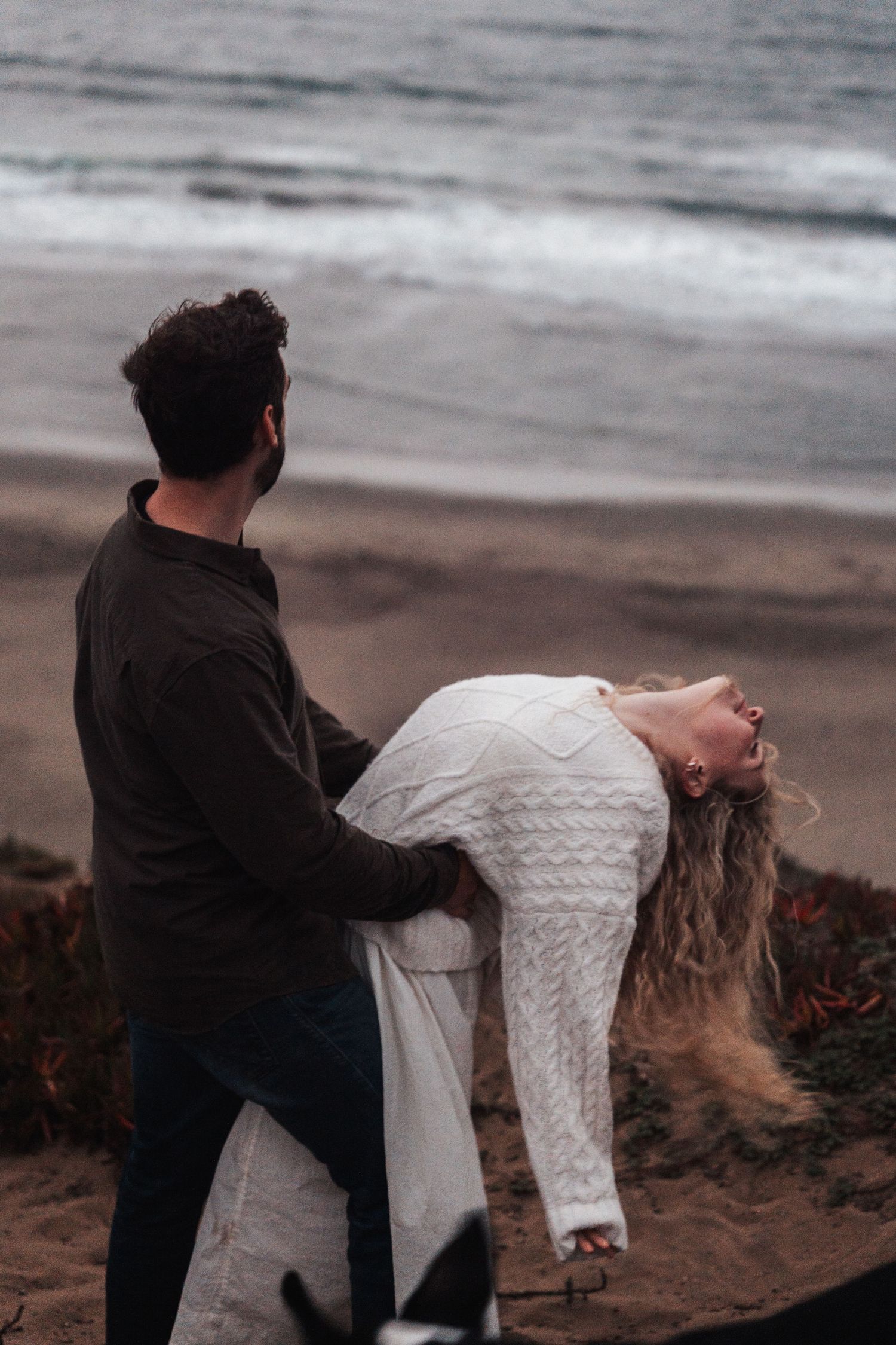 Couple sharing a romantic moment on a sandy beach, woman leaning back in man's arms near ocean waves.