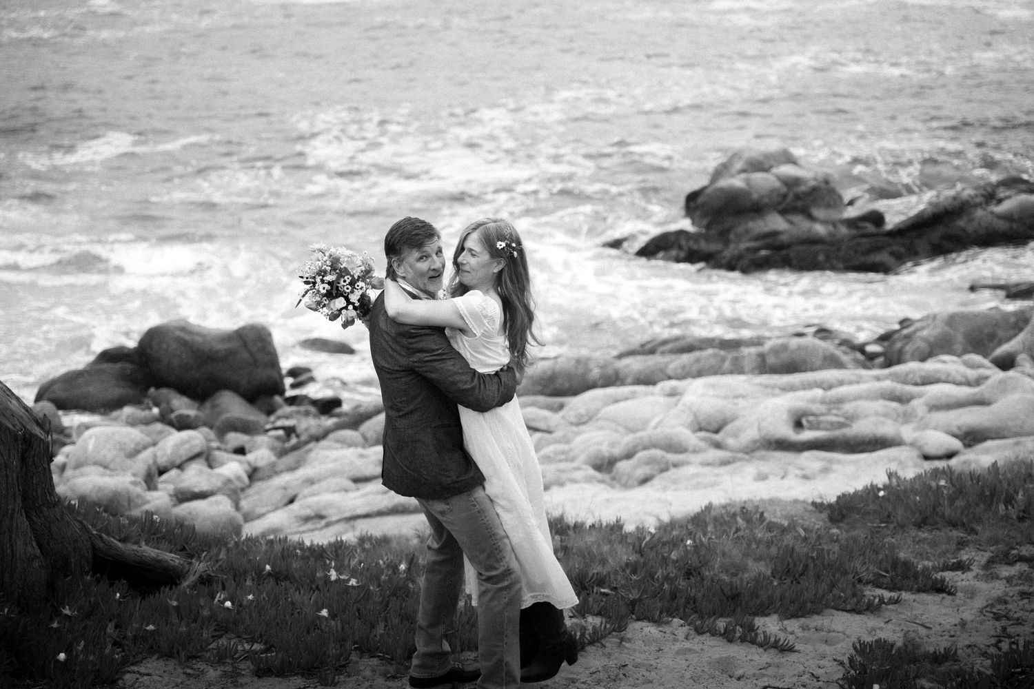 Black and white photo of a couple embracing on rocky coastal cliffs, bride holding bouquet with ocean waves behind them.