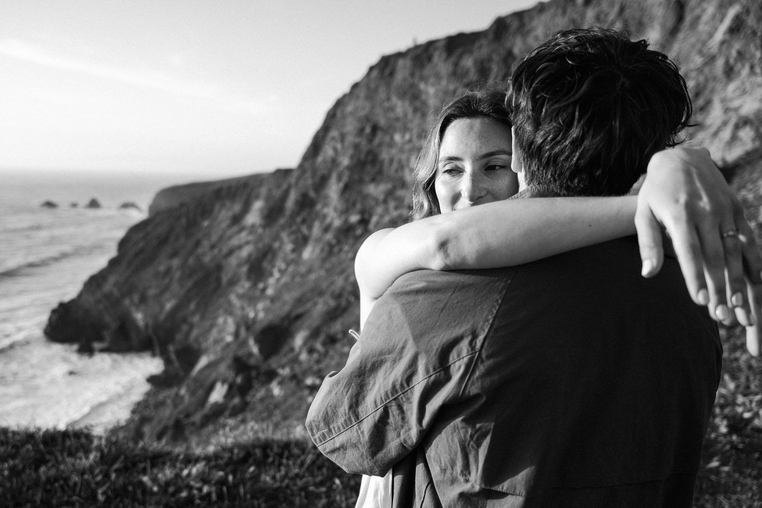 Black and white photo of couple embracing on dramatic ocean cliffs with rocky coastline in background.