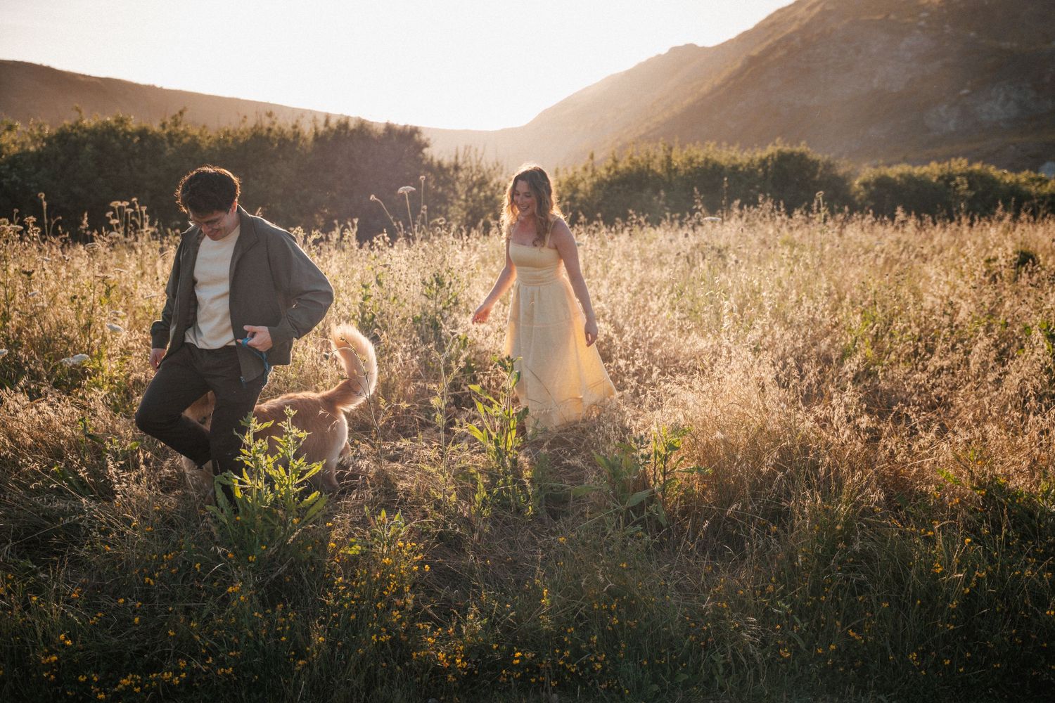 Couple playing with their dog in a golden sunlit field during sunset engagement photo session.