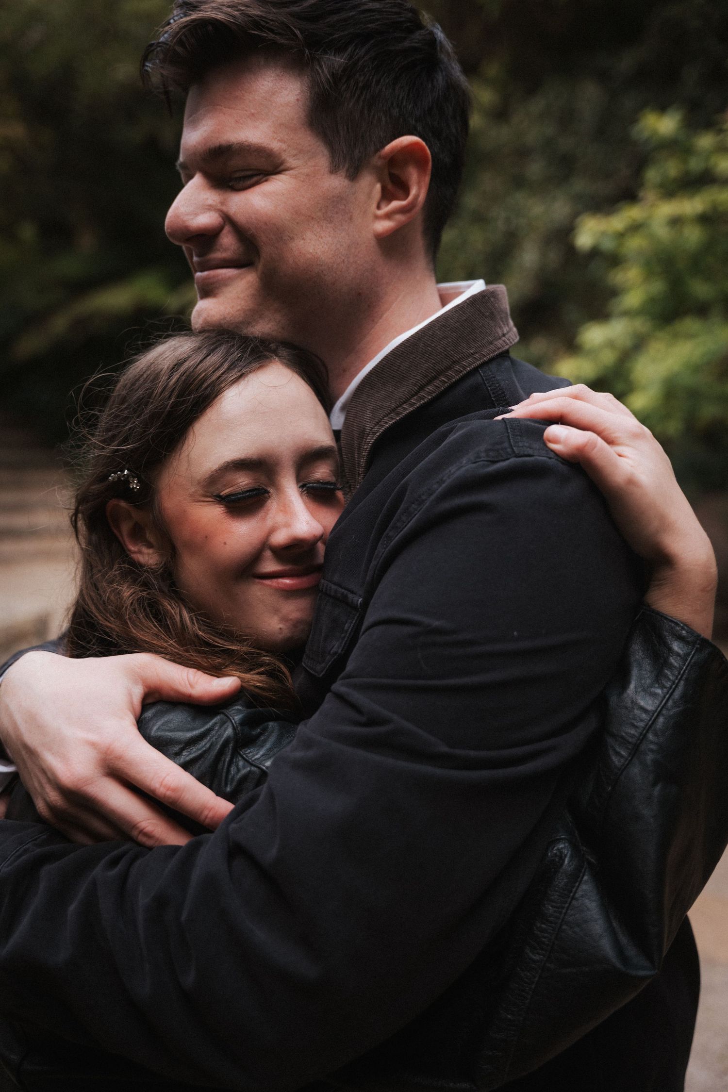 Happy couple sharing a tender hug outdoors, woman smiling with eyes closed in black jacket.