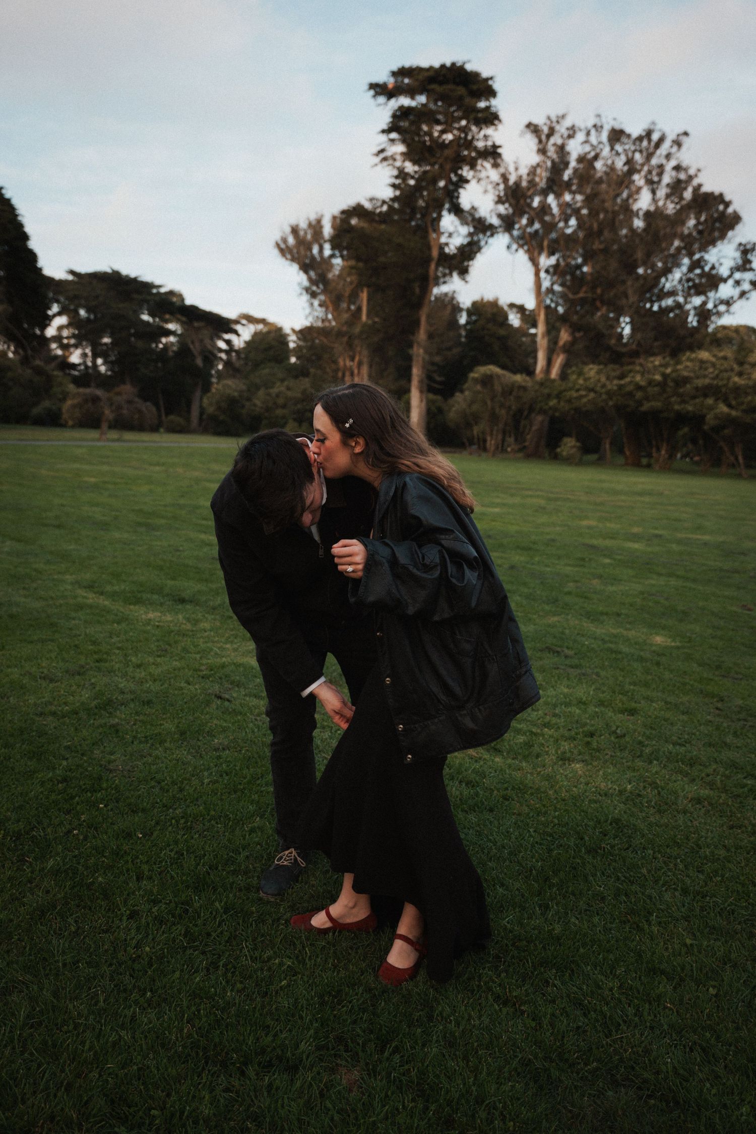 Two women dressed in black sharing a playful kiss on a green lawn at dusk.