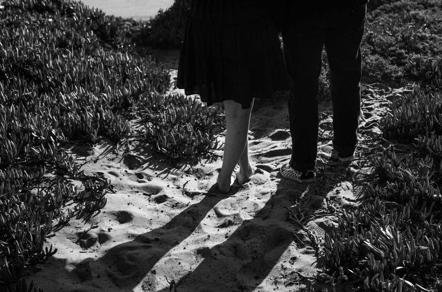 Black and white photo of a couple standing barefoot on sandy beach path surrounded by grass, casting long shadows.
