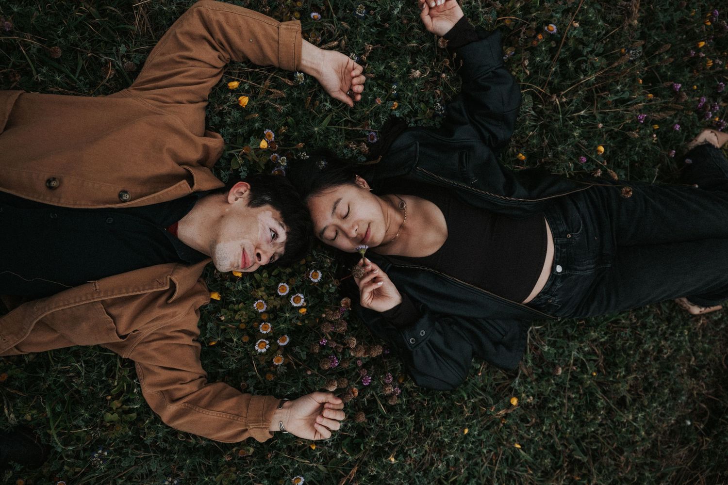 Two people lying on grass surrounded by wildflowers, eyes closed, in a peaceful and romantic outdoor setting.