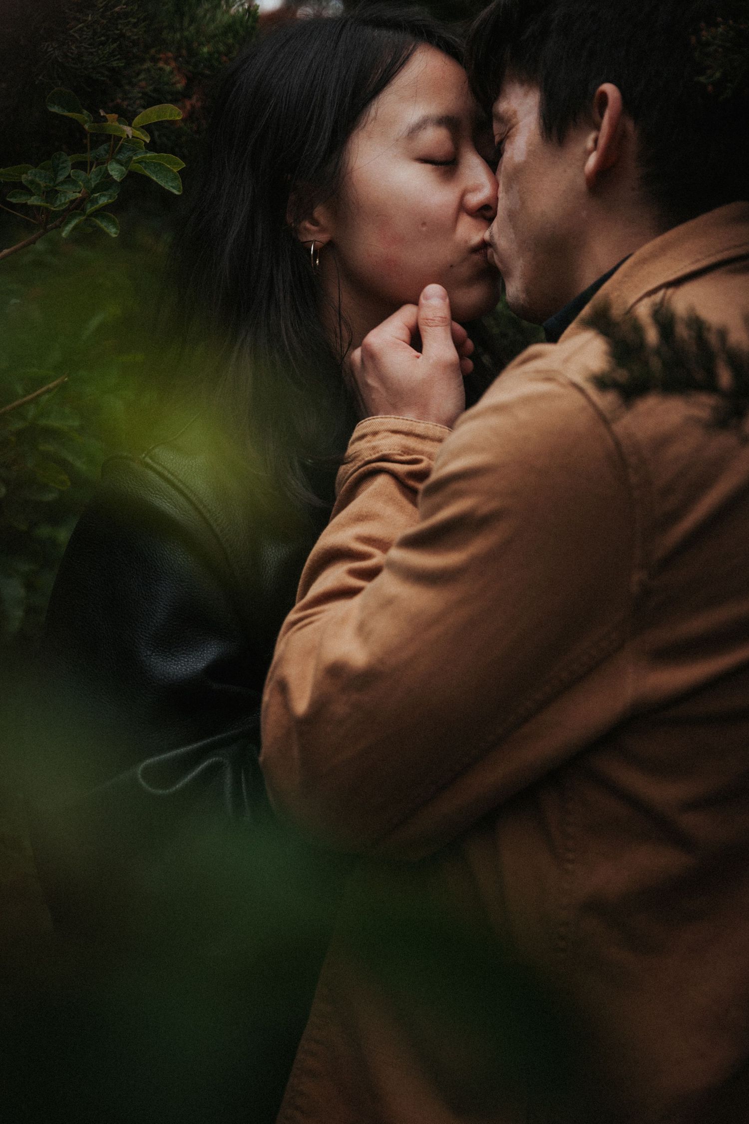 A couple shares a tender kiss surrounded by lush green foliage, the man wearing a brown jacket.