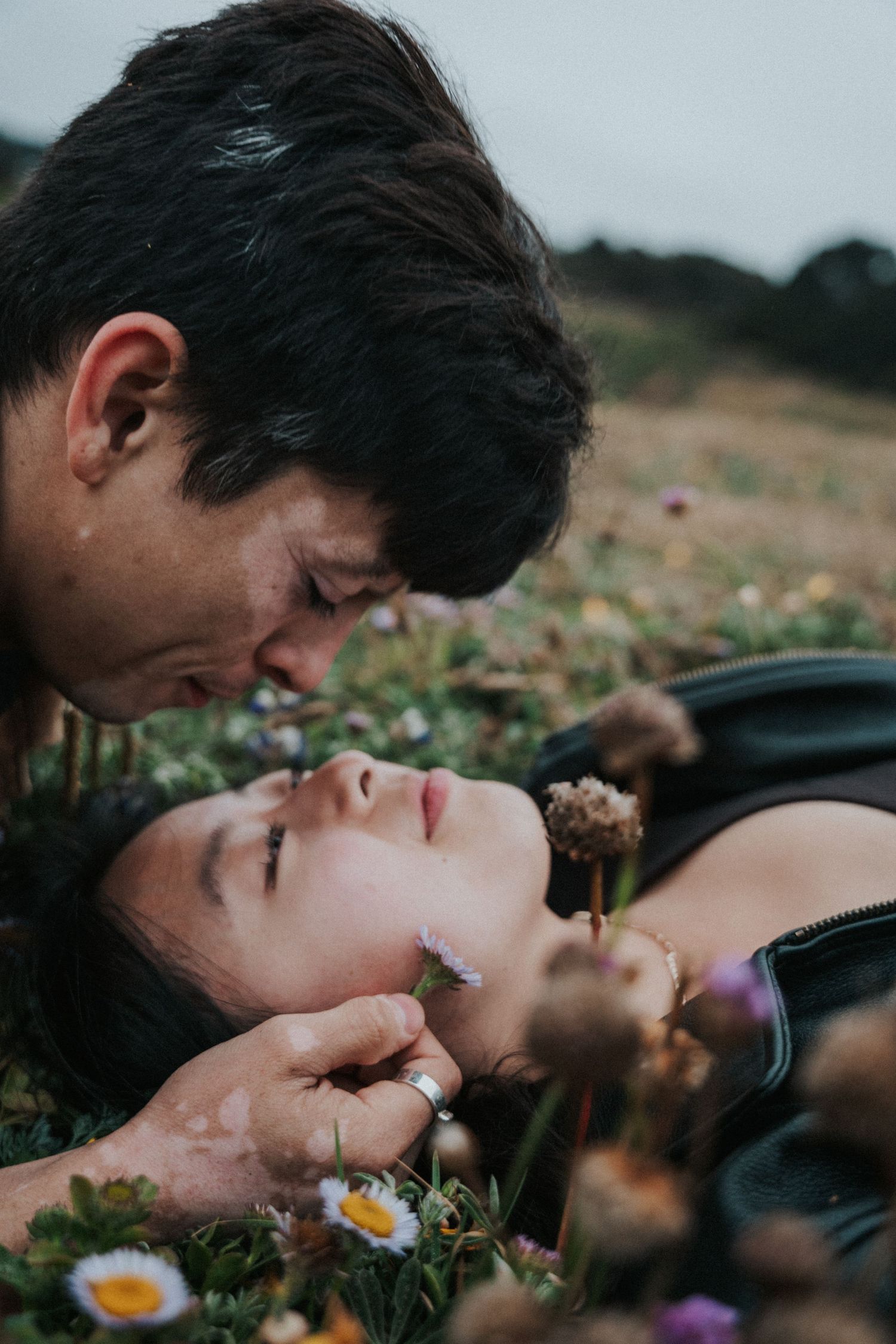 Romantic couple lying in a flower meadow, man leaning in to kiss woman holding a small wildflower.