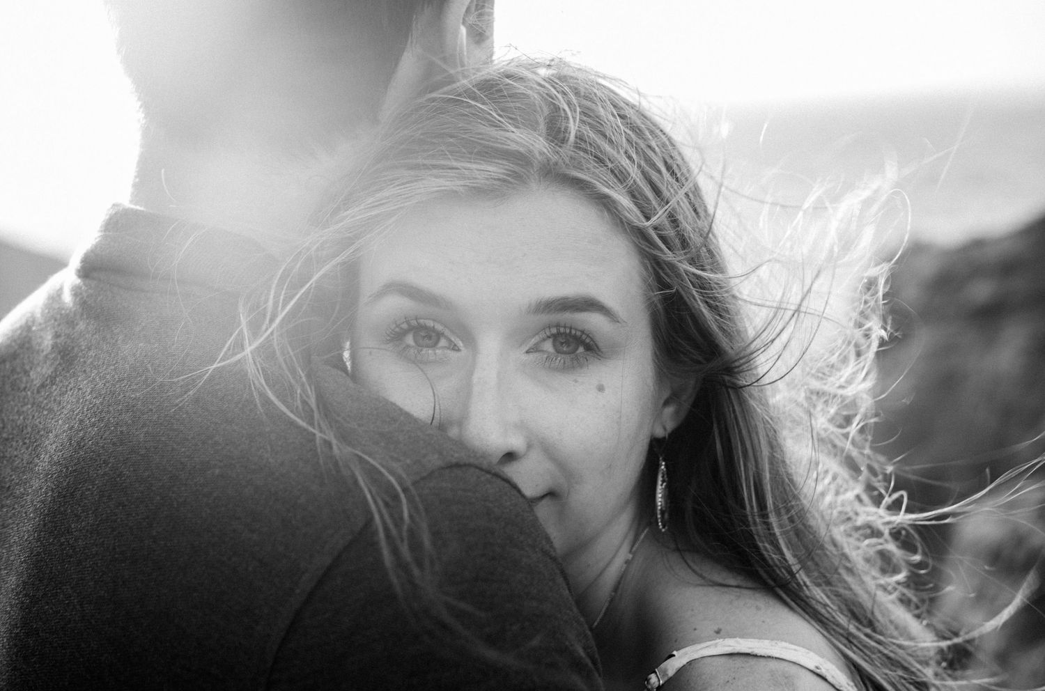 Black and white close-up of a woman resting her face on a mans shoulder, hair blowing in the wind.