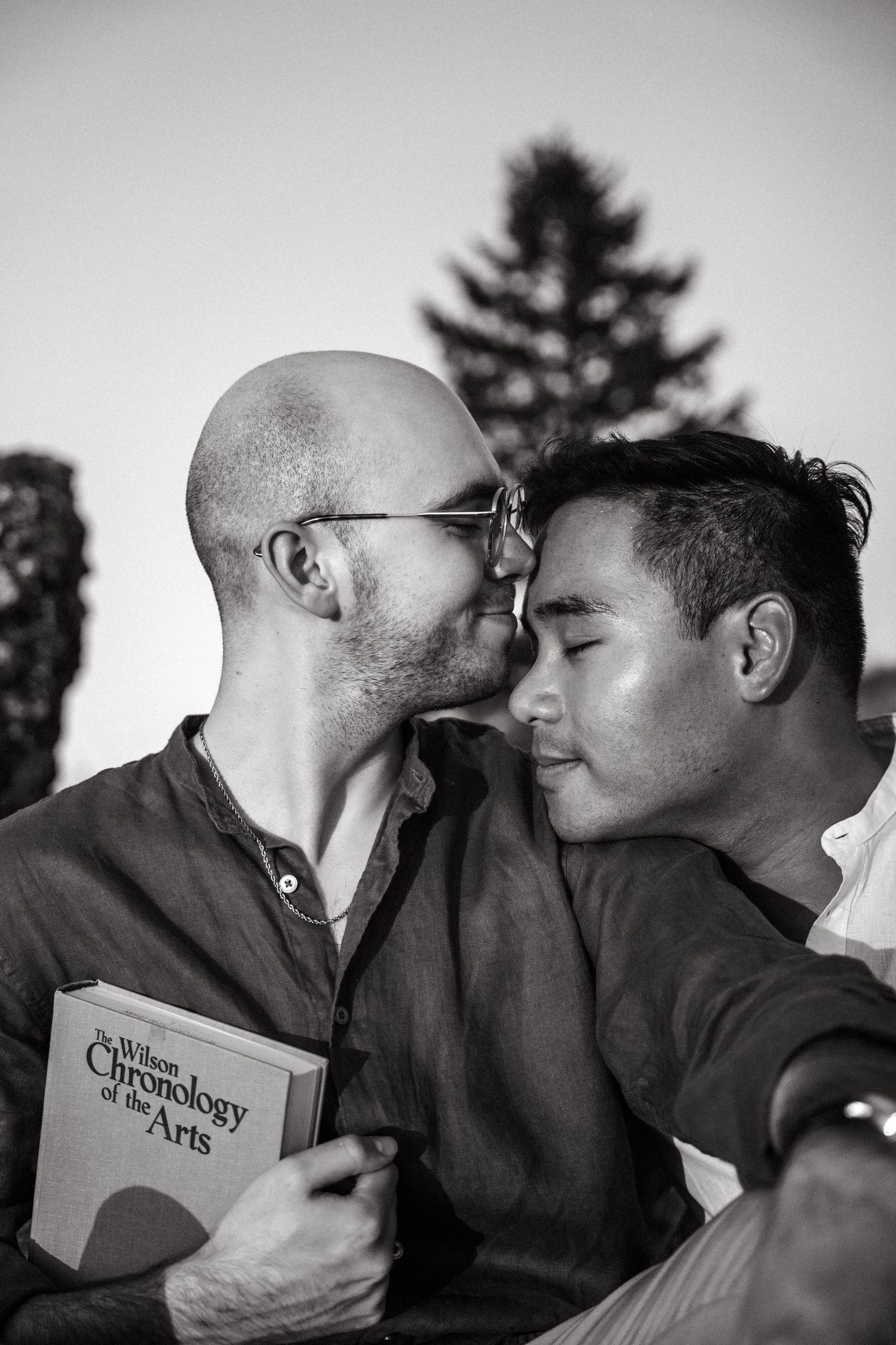 Two men share an intimate moment, nearly kissing, one holding a Chronology of the Arts book, in black and white.