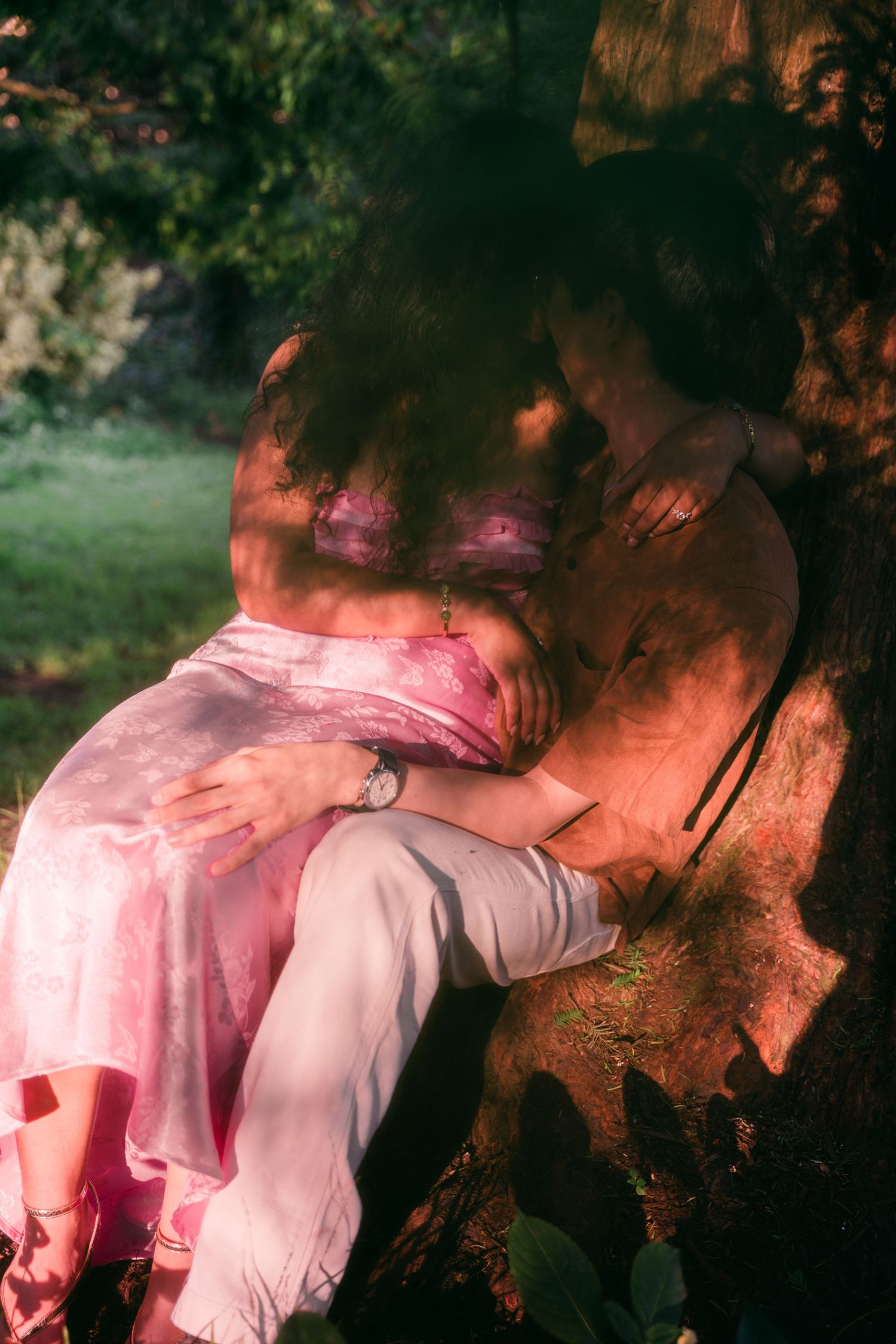 Couple embracing romantically against a tree in a sunlit garden, woman wearing a flowing pink dress.