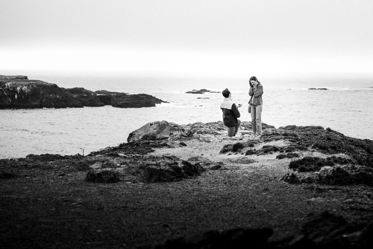 Black and white photo of a couple on rocky shoreline, one kneeling in a marriage proposal by the ocean.