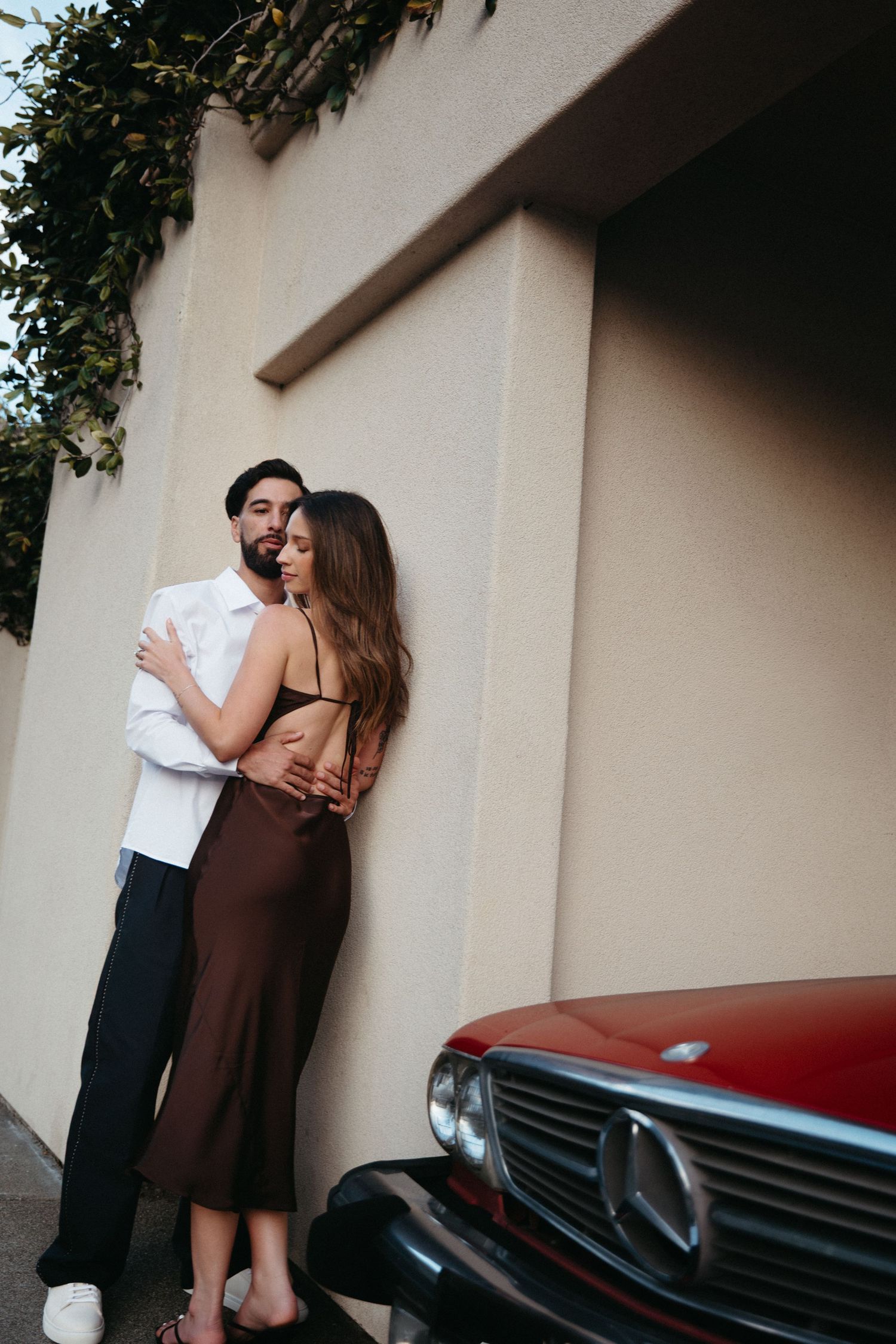A couple embraces romantically against a beige wall beside a vintage red Mercedes-Benz car.