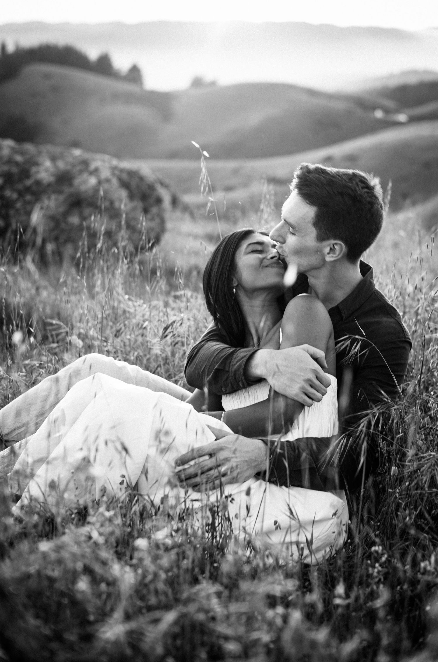 Black and white photo of a couple sitting in a meadow, sharing a romantic kiss surrounded by rolling hills.