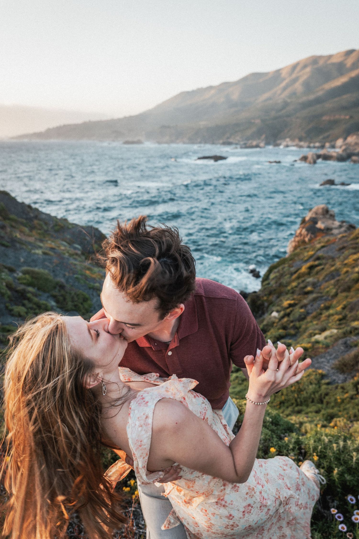 Man dipping woman for a kiss on rocky coastal cliffside during golden hour sunset.
