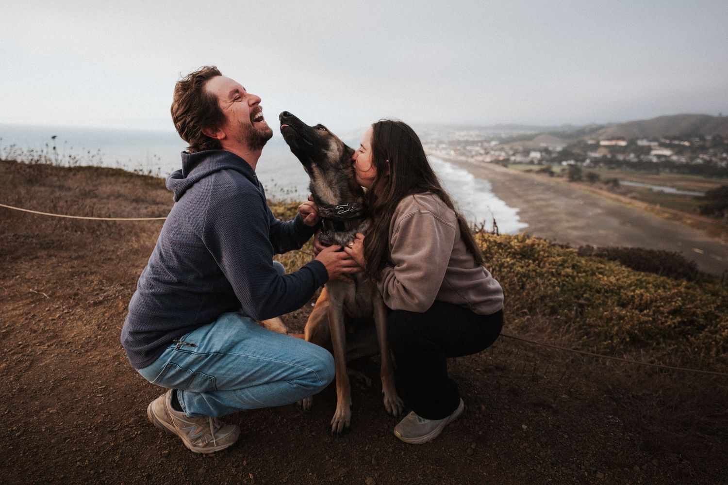 Couple crouching with their dog on a coastal hillside, sharing a joyful moment as the dog licks their faces.