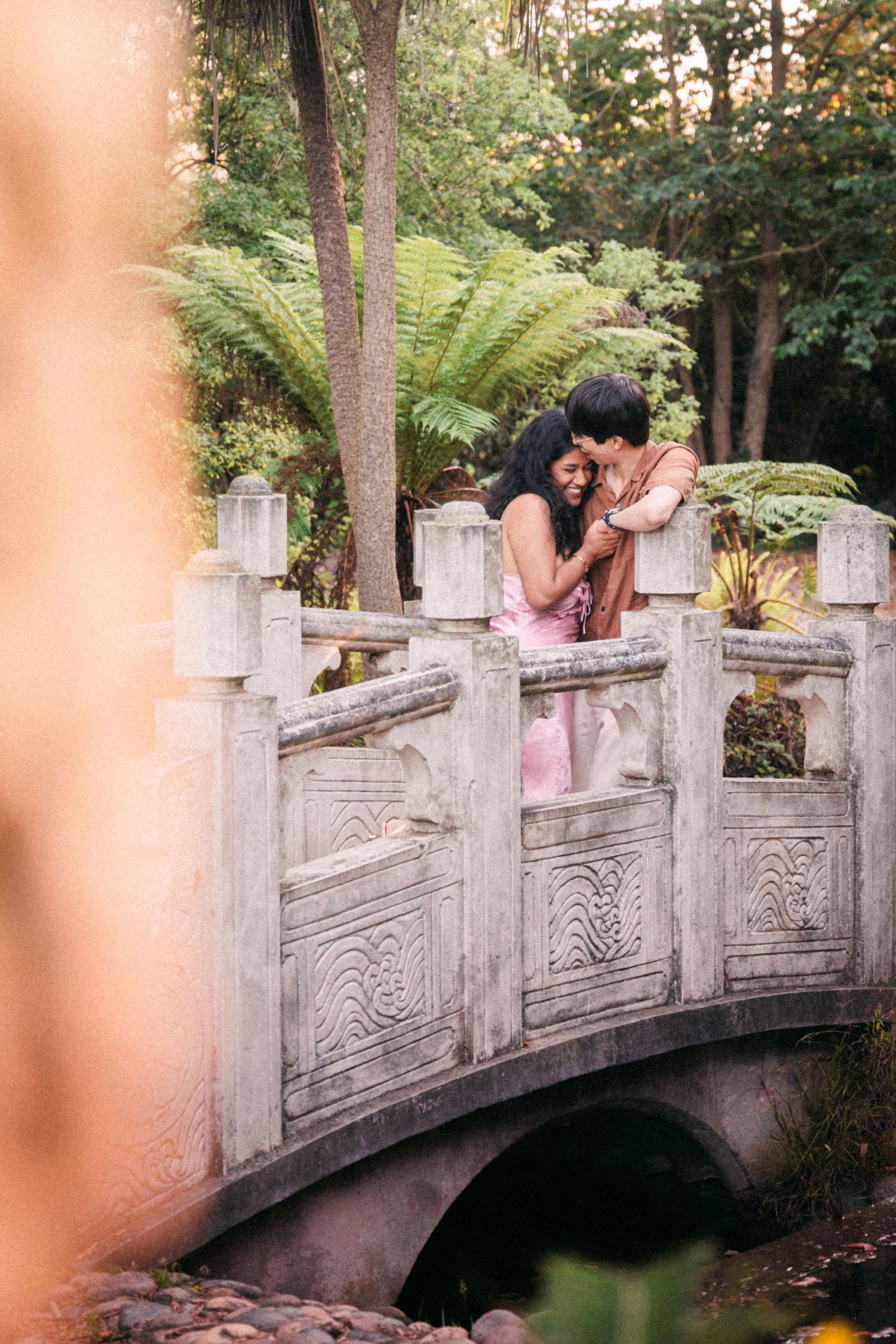 Couple sharing a tender embrace on an ornate stone bridge surrounded by tropical garden greenery.