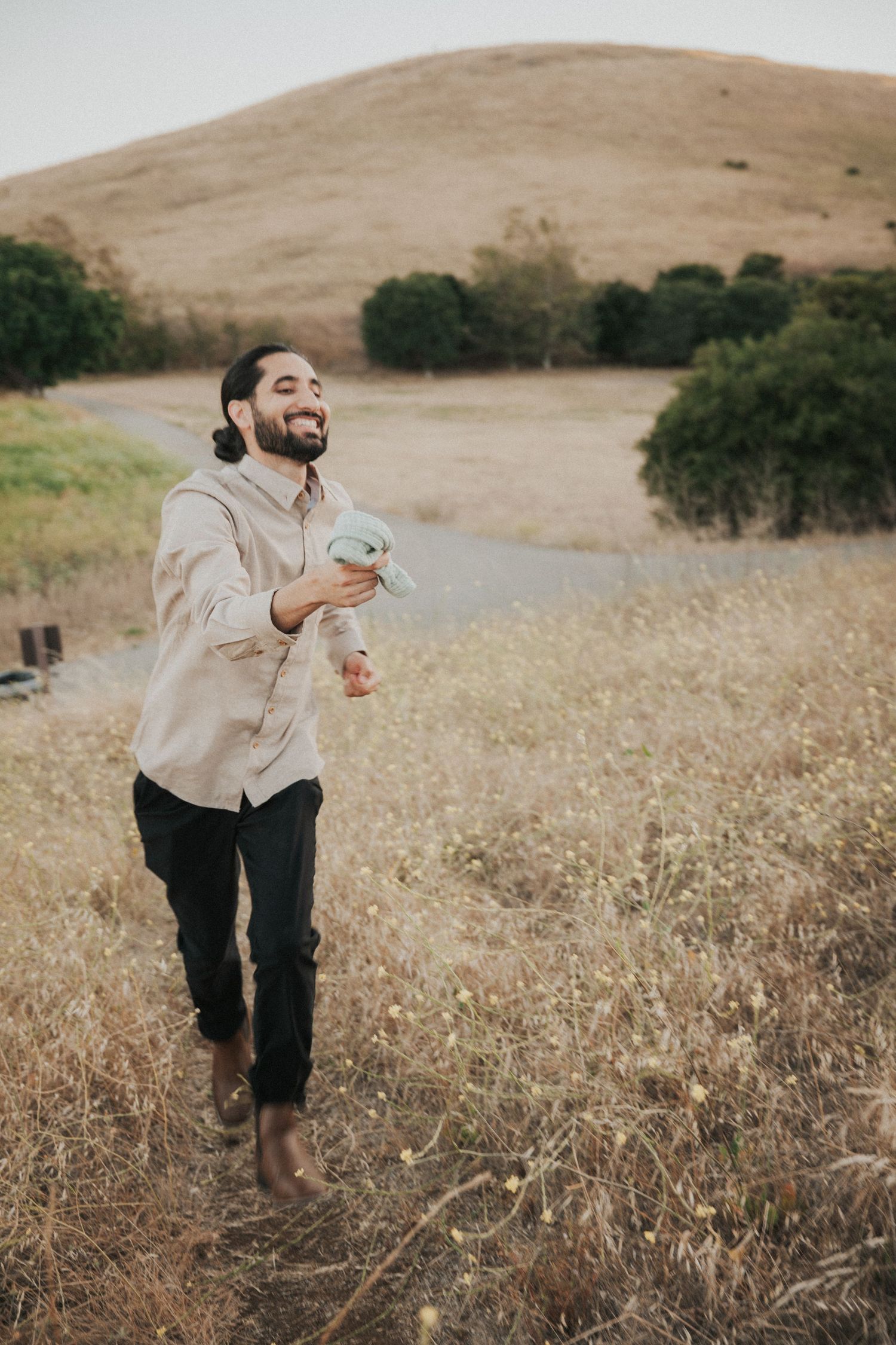 A bearded man with a bun laughs joyfully while walking barefoot through a golden meadow with rolling hills behind him.