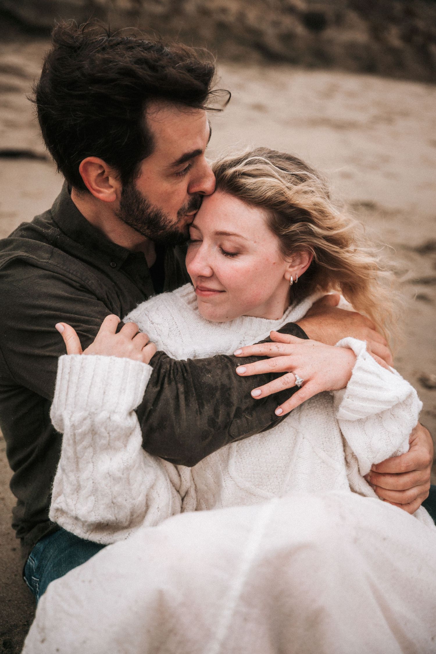 Man tenderly kissing woman on head while embracing on sandy beach on cloudy day.