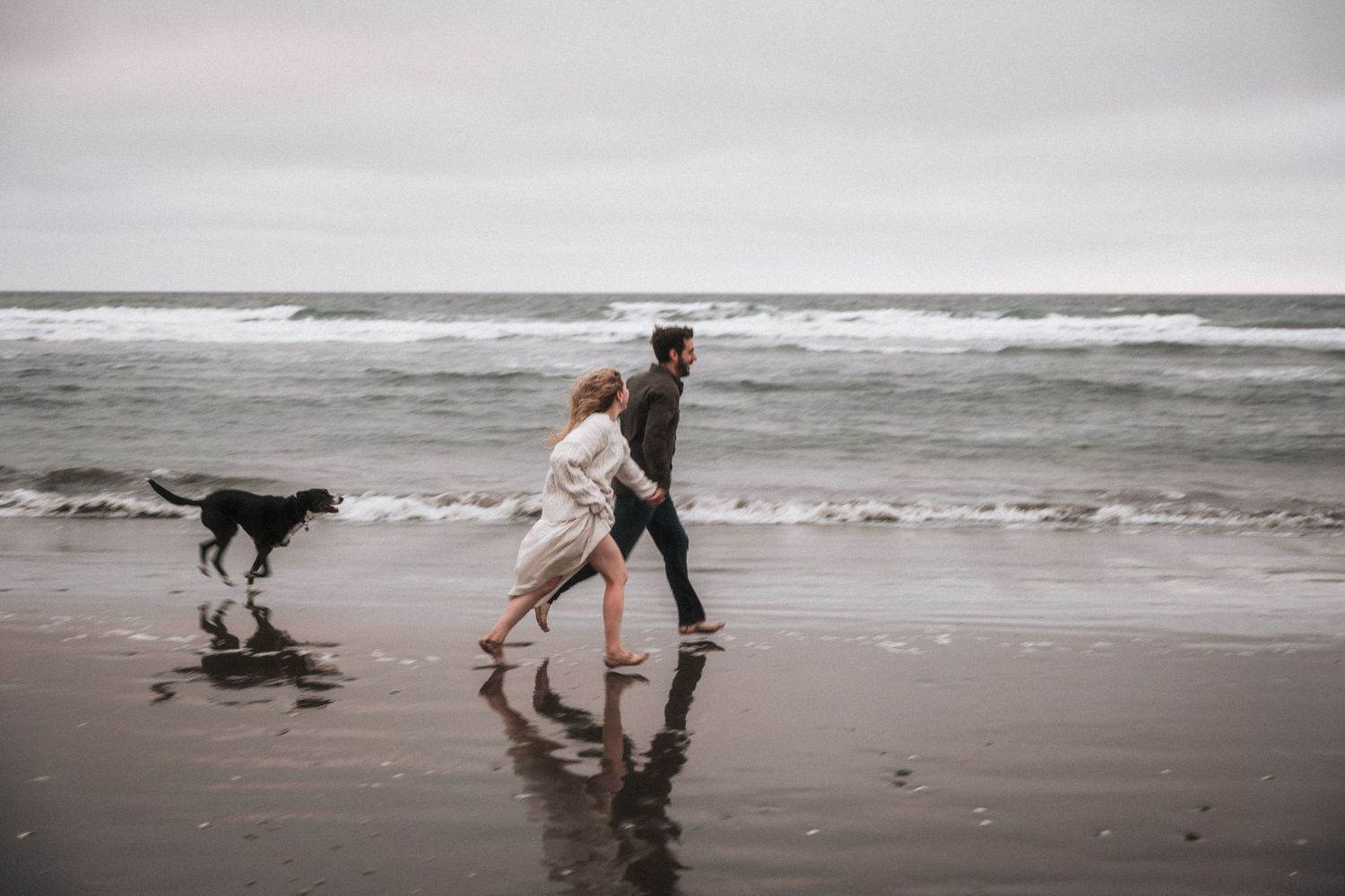 Couple walking barefoot on a wet beach with a black dog, ocean waves in the background on an overcast day.