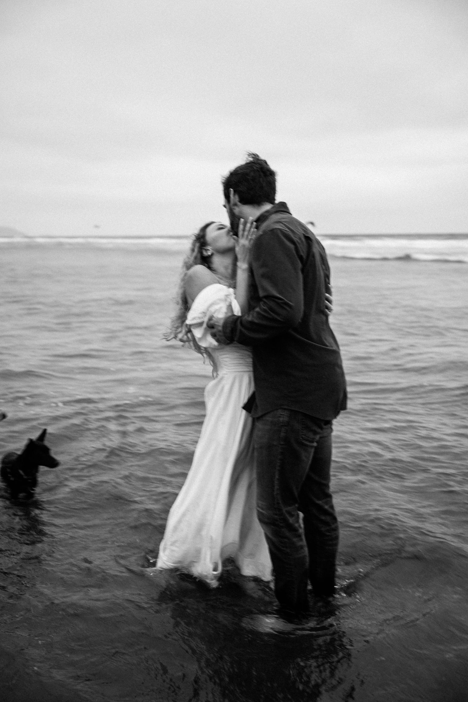 Black and white photo of a couple kissing in ocean waves, woman in white wedding dress, small dog nearby.