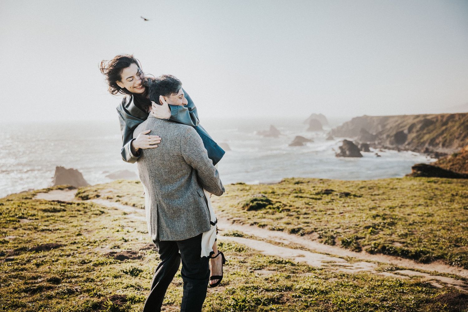 Couple embracing on a scenic coastal cliff with ocean rocks and clear sky in the background.