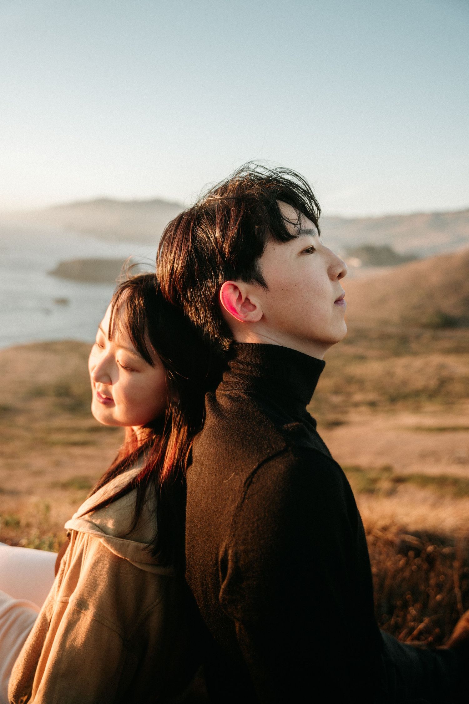 A man and woman stand back to back on a sunlit coastal bluff, gazing peacefully in opposite directions near the sea.