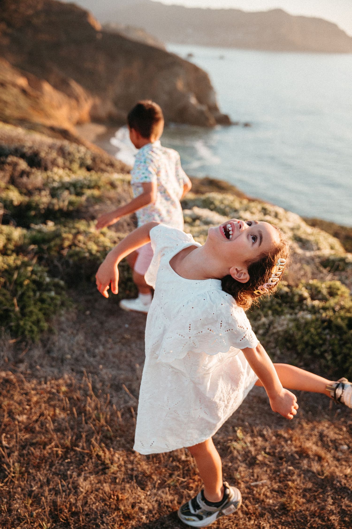 Two young girls in white dresses run and play on a coastal hillside at golden hour.