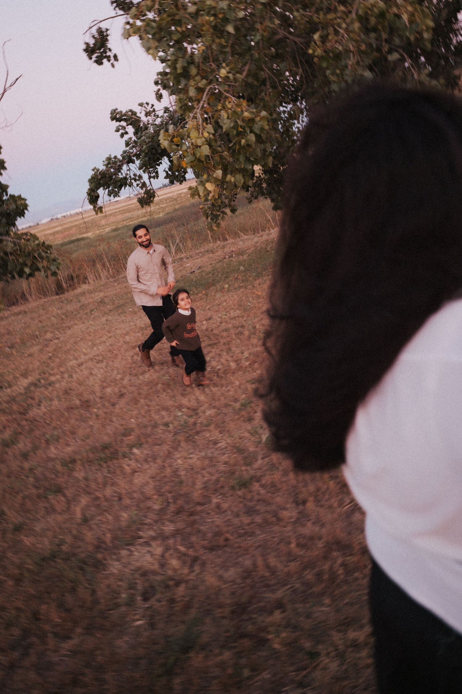 A father and young child run across a field toward a woman in the foreground at dusk.