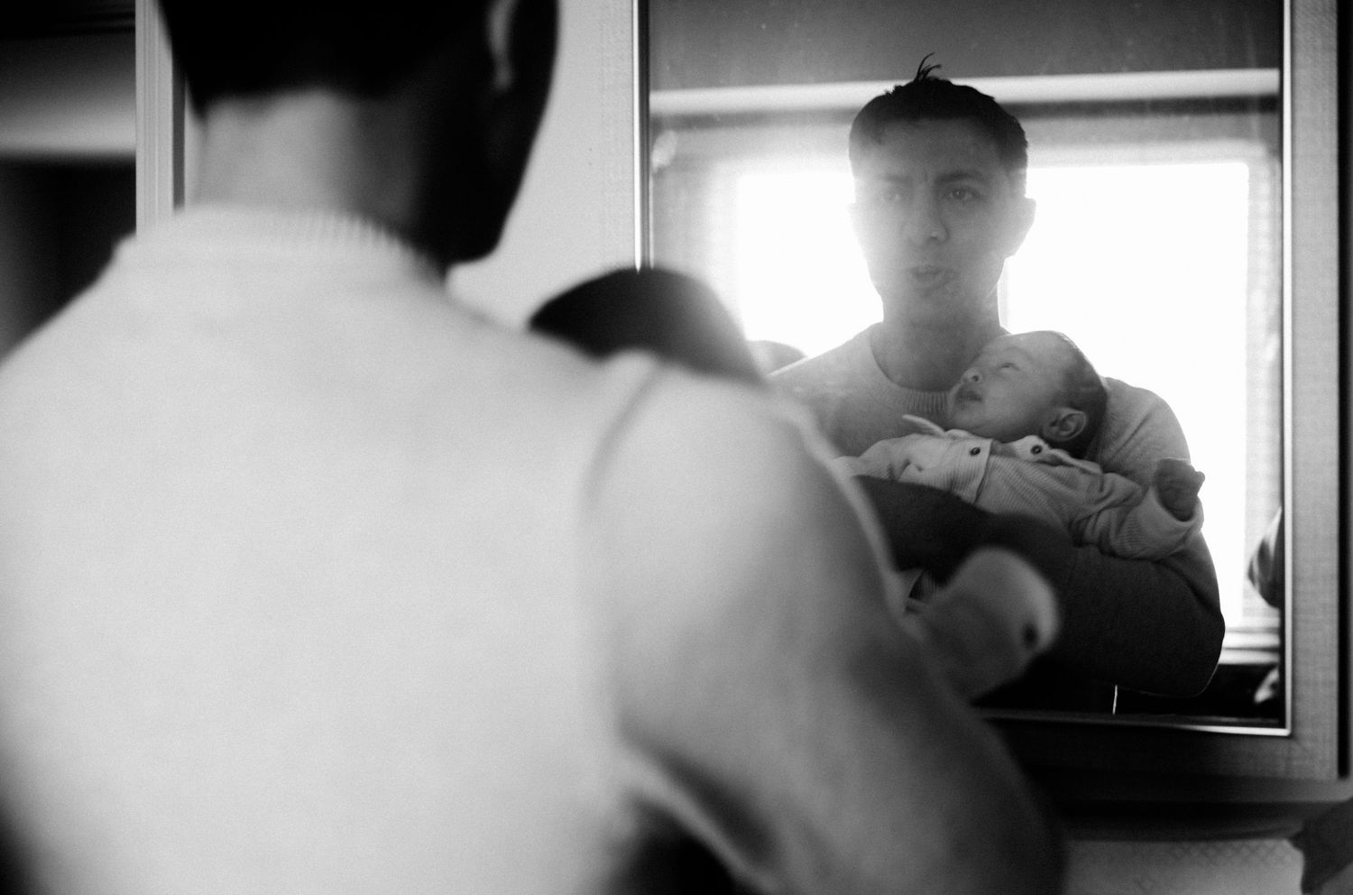 Black and white photo of a father holding his newborn baby, reflected in a mirror near a bright window.