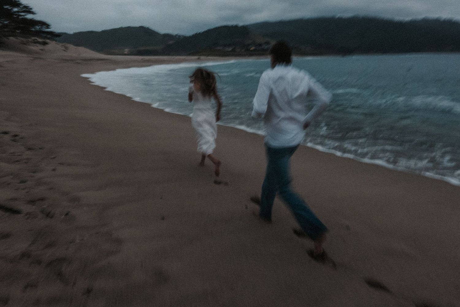 Couple in white outfits running barefoot along a sandy beach at dusk with ocean waves and misty mountains behind.