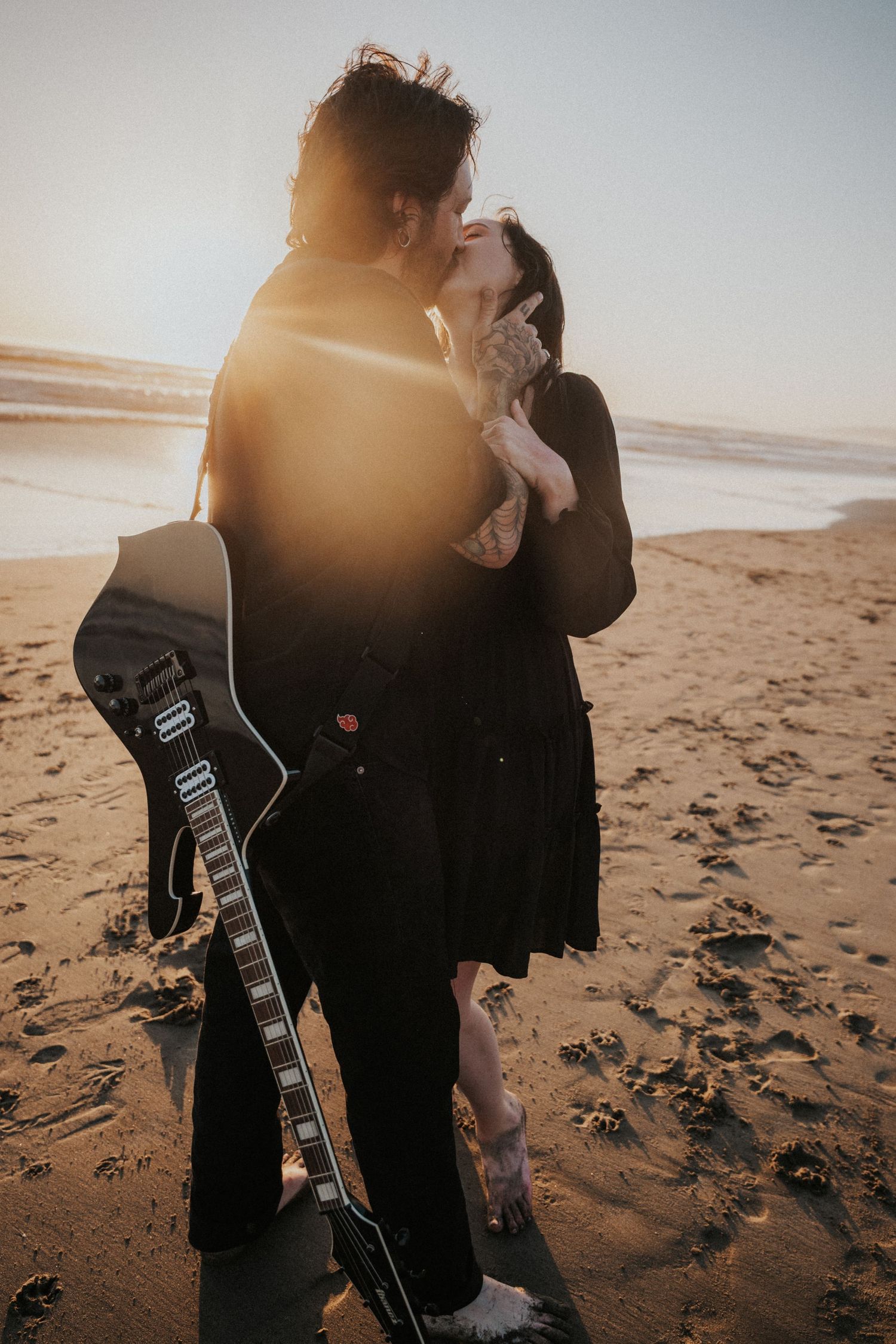 Couple kissing on beach at sunset with black electric guitar, wearing dark clothing, ocean waves in background. Photography by The Y.K. Stud