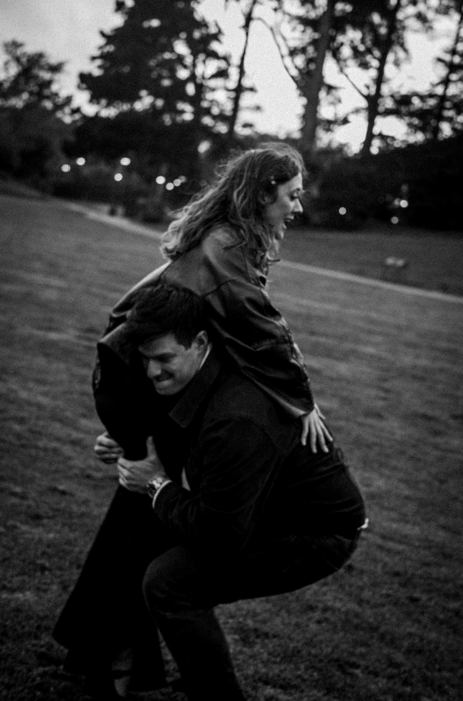 Black and white photo of a man giving a laughing woman a piggyback ride on a grassy field.