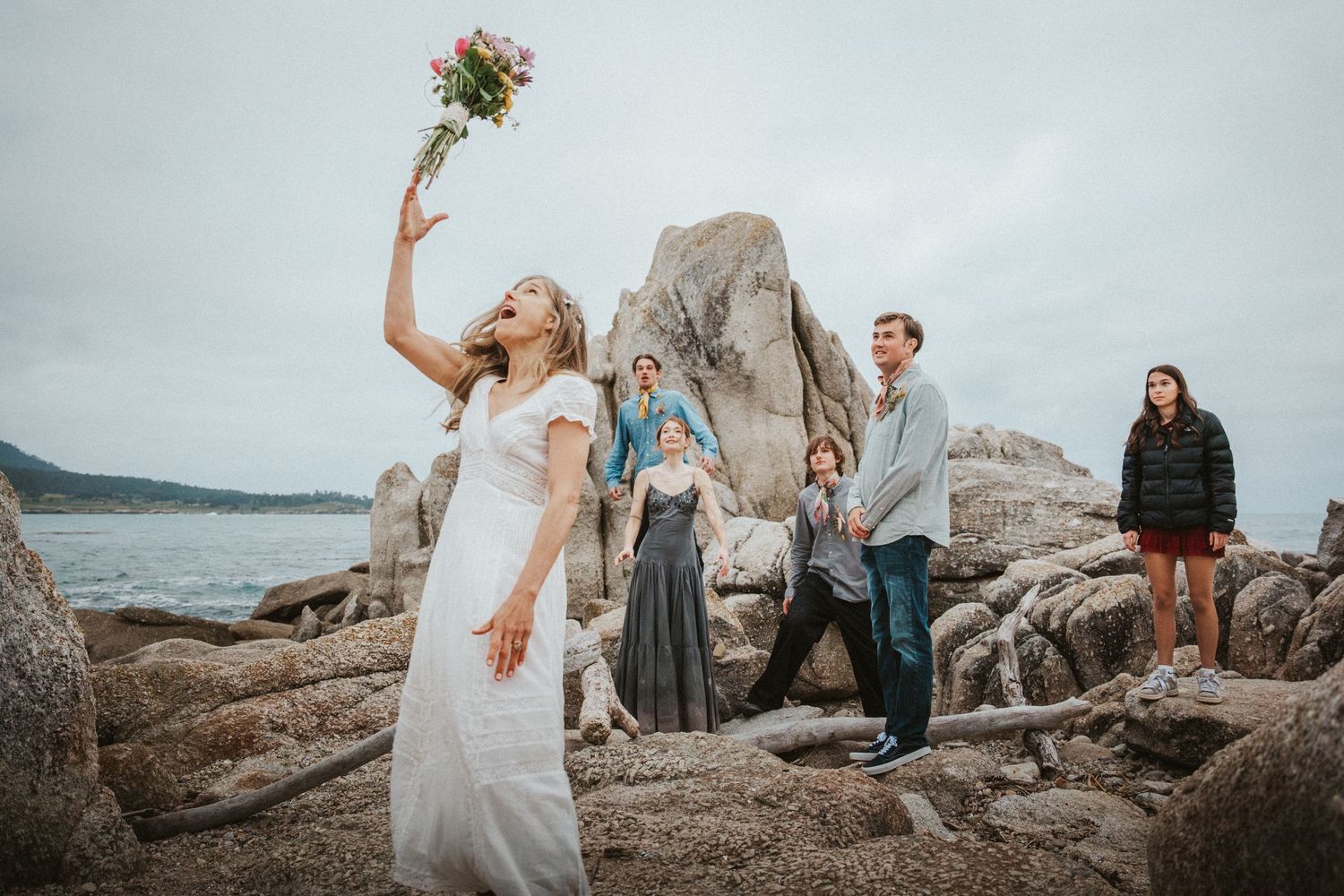 A bride tosses her bouquet to guests standing on rocky coastal cliffs during a wedding celebration.