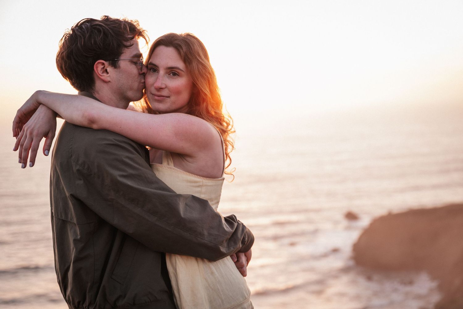 Romantic couple embracing at sunset by the ocean, woman smiling as man kisses her cheek.