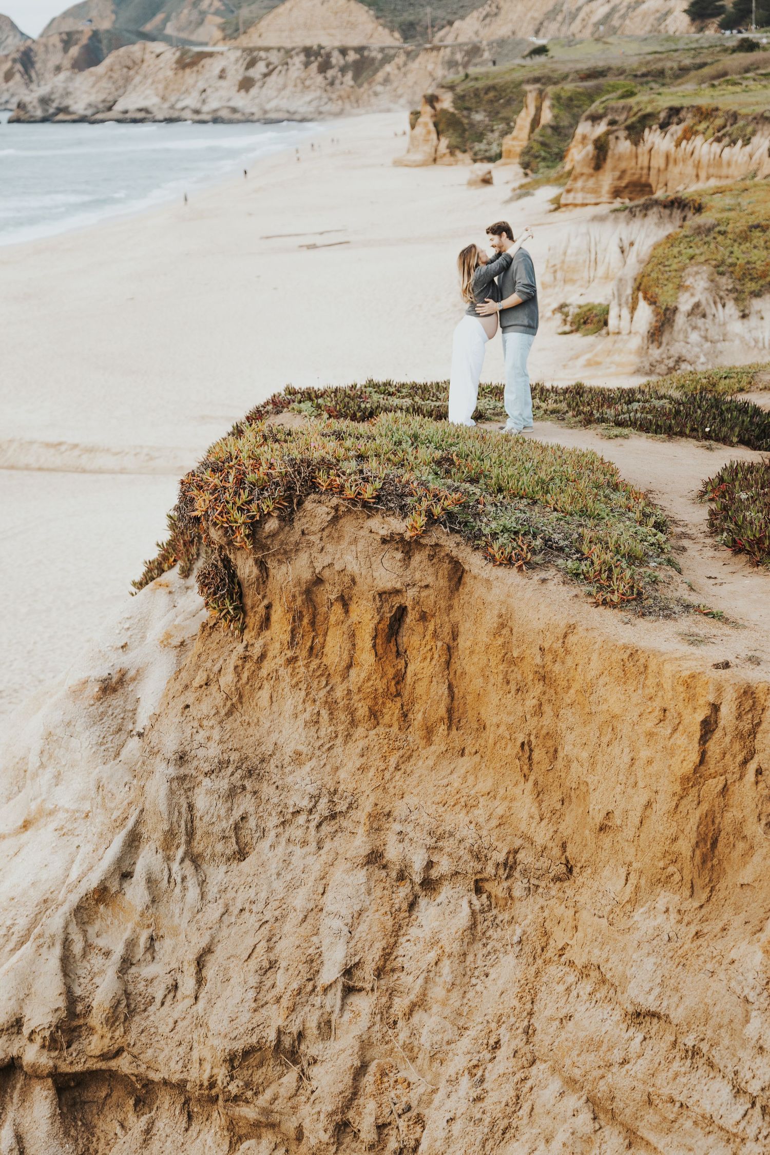 Couple embracing on a sandy coastal cliff overlooking a scenic beach with golden cliffs and ocean waves in the background.