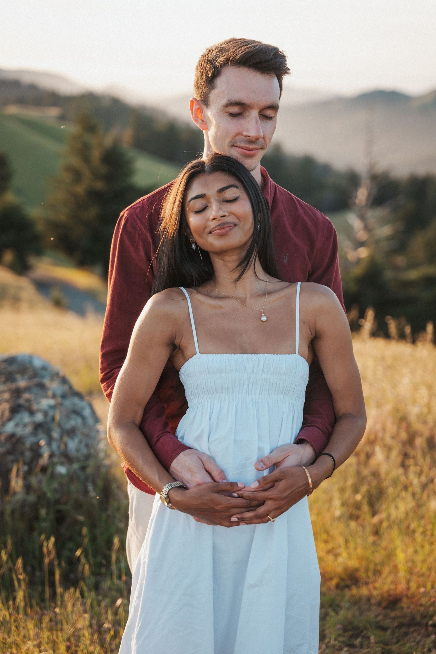 Couple embracing in golden field at sunset, woman in white dress, man in burgundy shirt, rolling hills background.
