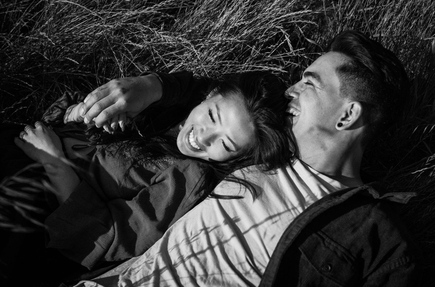 Black and white photo of a laughing couple lying in grass, sharing a joyful moment together.Photography by The Y.K. Studio.