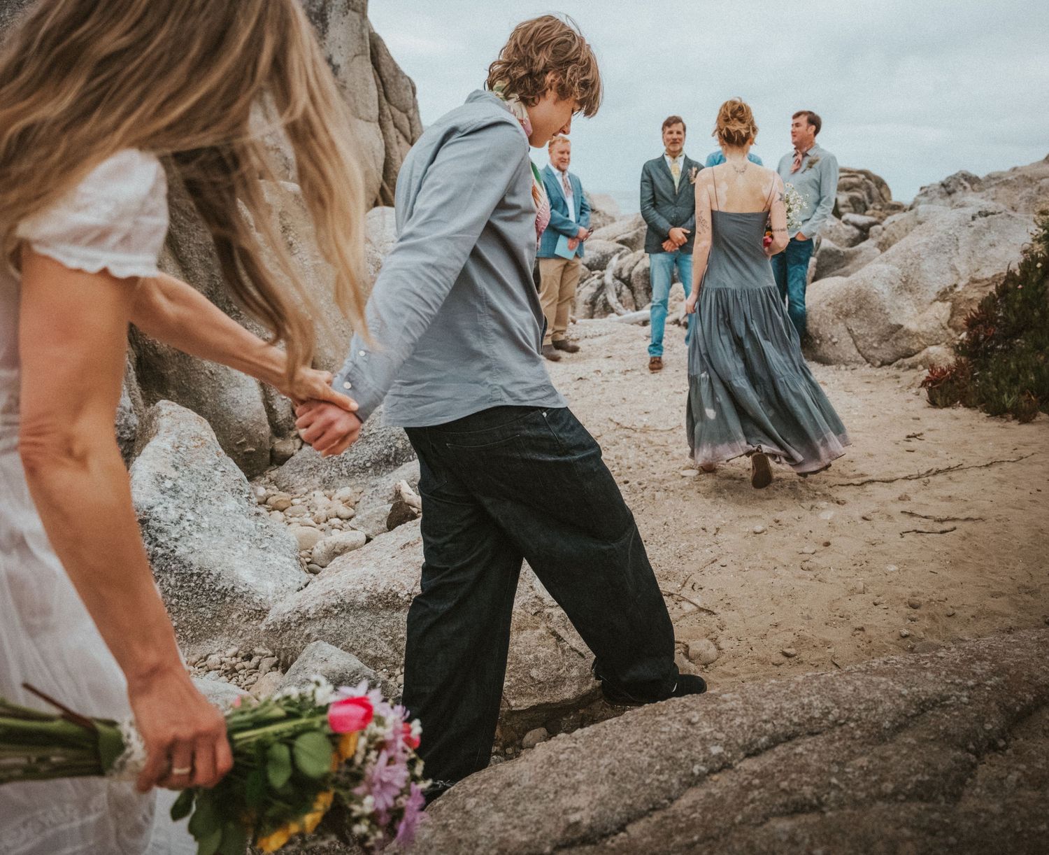 Wedding party walking on sandy beach with large rocks, bride holding colorful bouquet.