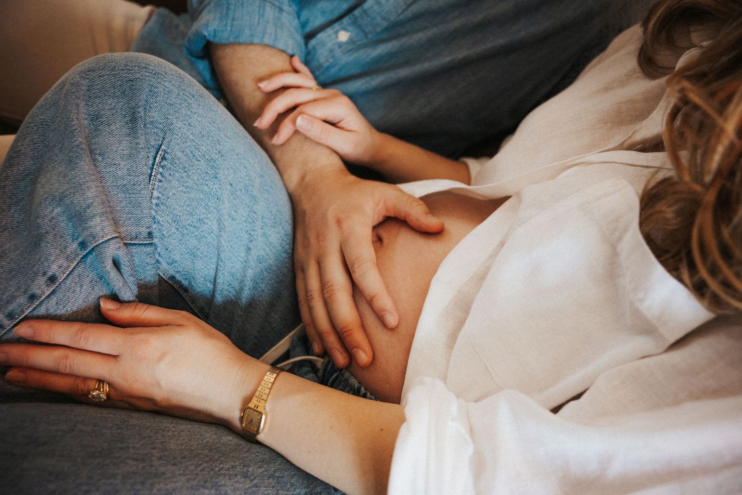 Couple relaxing on sofa, hands tenderly touching pregnant woman's belly, wearing jeans and white shirt.