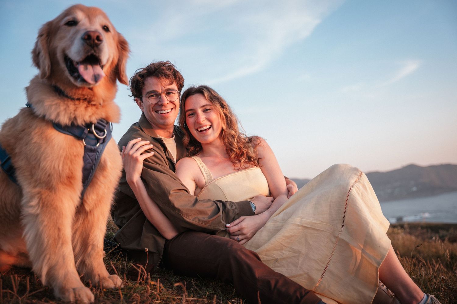 Smiling couple relaxing in grass with happy golden retriever dog under a blue sky.